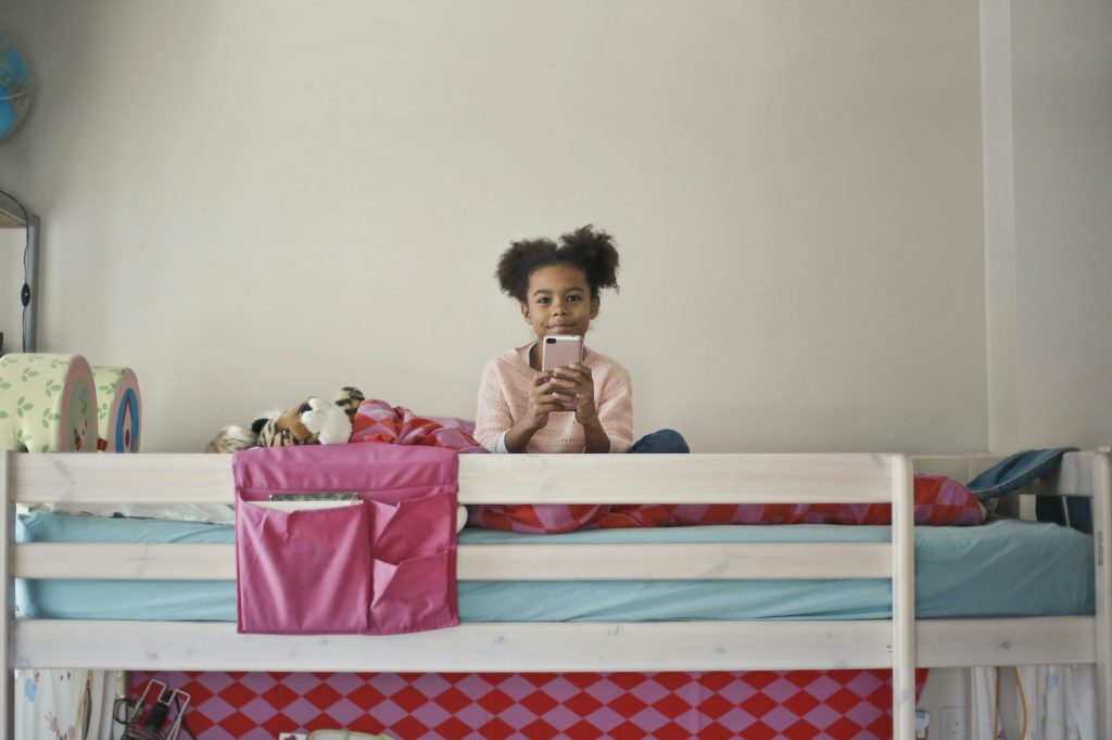 The image shows a young girl sitting on her bed, using a smartphone and smiling. Credit: bruce mars on Unsplash