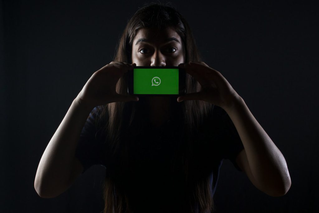 Girl standing in the dark, holding a phone sideways, facing the photographer directly. On the display there's a whatsapp icon. The image represents the dangers of whatsapp.